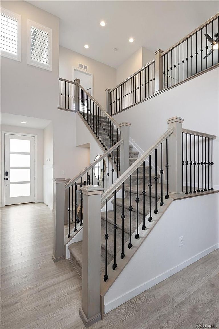 This bright and airy entryway features a grand staircase with light-toned wooden handrails and elegant black metal balusters. The space is finished with neutral wall colors and light wood-look flooring, creating a clean, modern aesthetic. The perspective captures the open, multi-level layout, emphasizing the architectural flow and welcoming atmosphere of the home.