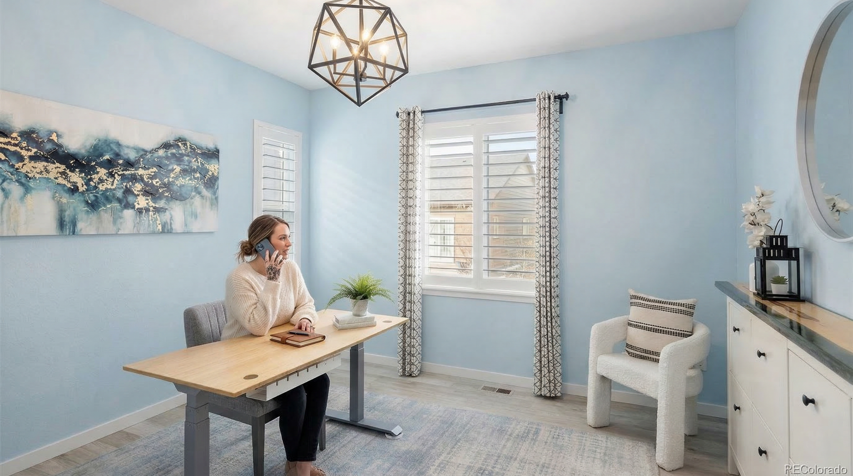 This bright home office features light blue walls, a contemporary light wood standing desk, and an elegant geometric pendant chandelier. A cozy white textured armchair and a framed abstract painting complete the inviting and productive workspace. The room is well-lit with natural light filtering through shuttered windows, creating a clean and professional atmosphere.