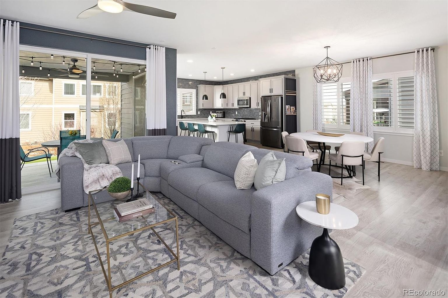 This open-concept living space features a large, plush grey sectional sofa as the centerpiece, set atop a patterned area rug. The room flows seamlessly into a bright dining area and a modern kitchen with white cabinetry and a central island, all illuminated by contemporary lighting fixtures. Large sliding glass doors provide a view of the patio, creating an airy and inviting atmosphere perfect for modern living.