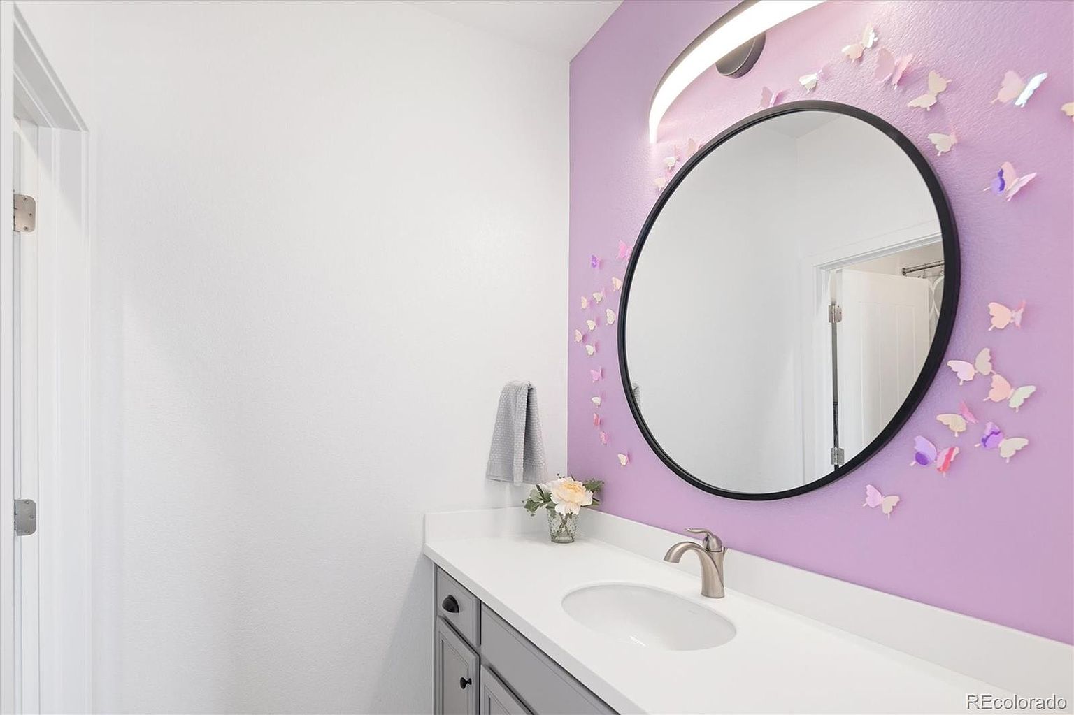 This charming bathroom features a vibrant purple accent wall adorned with decorative 3D butterflies, creating a whimsical and playful aesthetic. The space includes a sleek, modern circular mirror, a white vanity with a gray base, and a polished chrome faucet. The clean white countertop and neutral surrounding walls balance the bold color, making it an inviting and unique space.