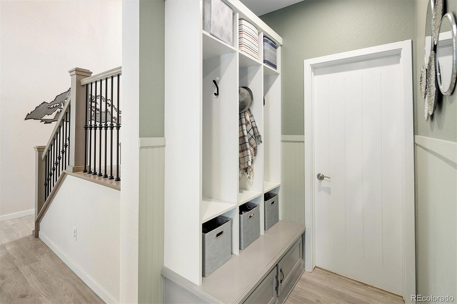 This interior view captures a functional entryway featuring a built-in mudroom bench with storage cubbies and hooks, accented by a soft sage green wall color and white wainscoting. To the left, a staircase with dark metal balusters and a decorative mountain-themed wall art piece adds a touch of character to the space. The perspective is eye-level, highlighting the clean, organized transition area that leads toward a closed white door.