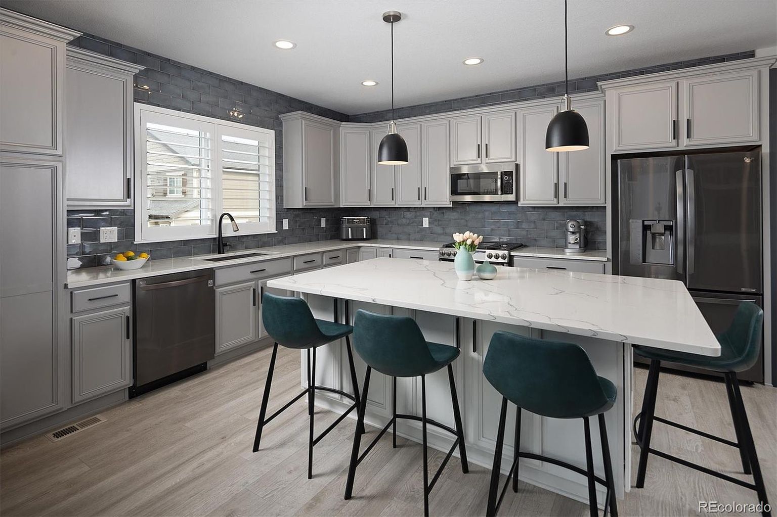 This modern, well-lit kitchen features light gray cabinetry paired with a striking dark blue subway tile backsplash and white quartz countertops. A large central island serves as a focal point, complemented by three teal bar stools and sleek black pendant lighting. The space is equipped with stainless steel appliances, including a refrigerator and microwave, creating a clean and contemporary aesthetic.