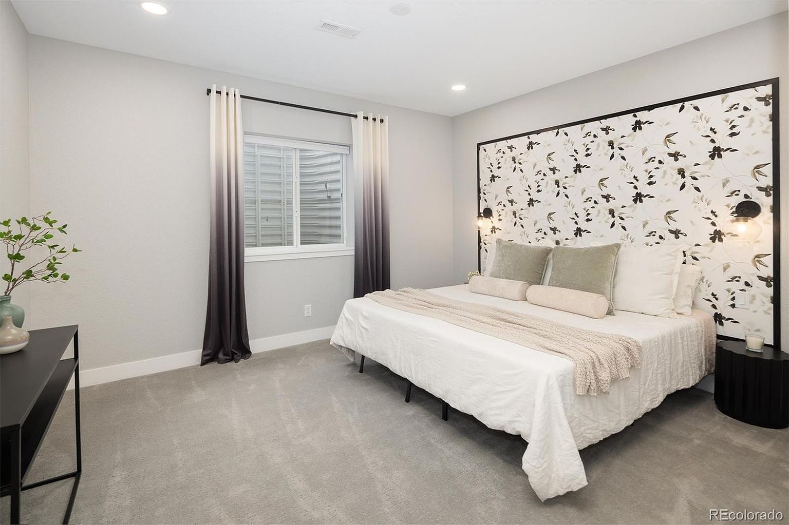 This spacious primary bedroom features a striking floral-patterned accent wall behind a large bed dressed in crisp white linens and neutral throw pillows. The room is anchored by a plush gray carpet and illuminated by modern wall-mounted sconces, while floor-to-ceiling ombre curtains frame a window looking out to a window well. The overall aesthetic is contemporary and serene, offering a clean, inviting atmosphere.
