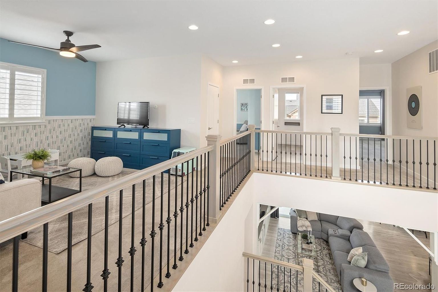 This elevated perspective captures a bright, open-concept loft area overlooking a lower-level living space. The upper level features a cozy lounge area with a blue dresser, a ceiling fan, and a neutral color palette, while the black metal railing provides a modern architectural contrast. The view highlights the home's airy, multi-level design and seamless transition between the upstairs landing and the main floor living room.