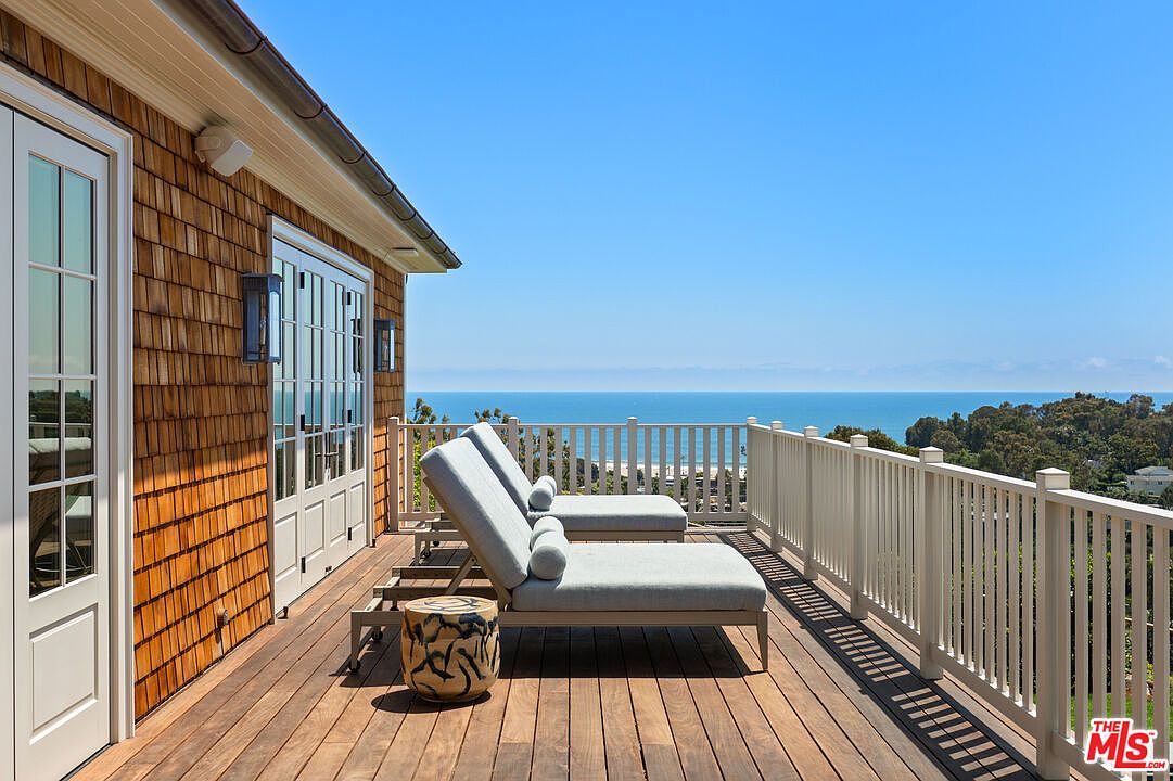 This image showcases a beautiful outdoor deck area with stunning ocean views. The deck features wooden flooring, white railings, and comfortable lounge chairs, creating a relaxing and inviting space. The architectural style of the house is visible with its shingle siding and white-framed windows and doors, adding to the overall charm and appeal of the property.