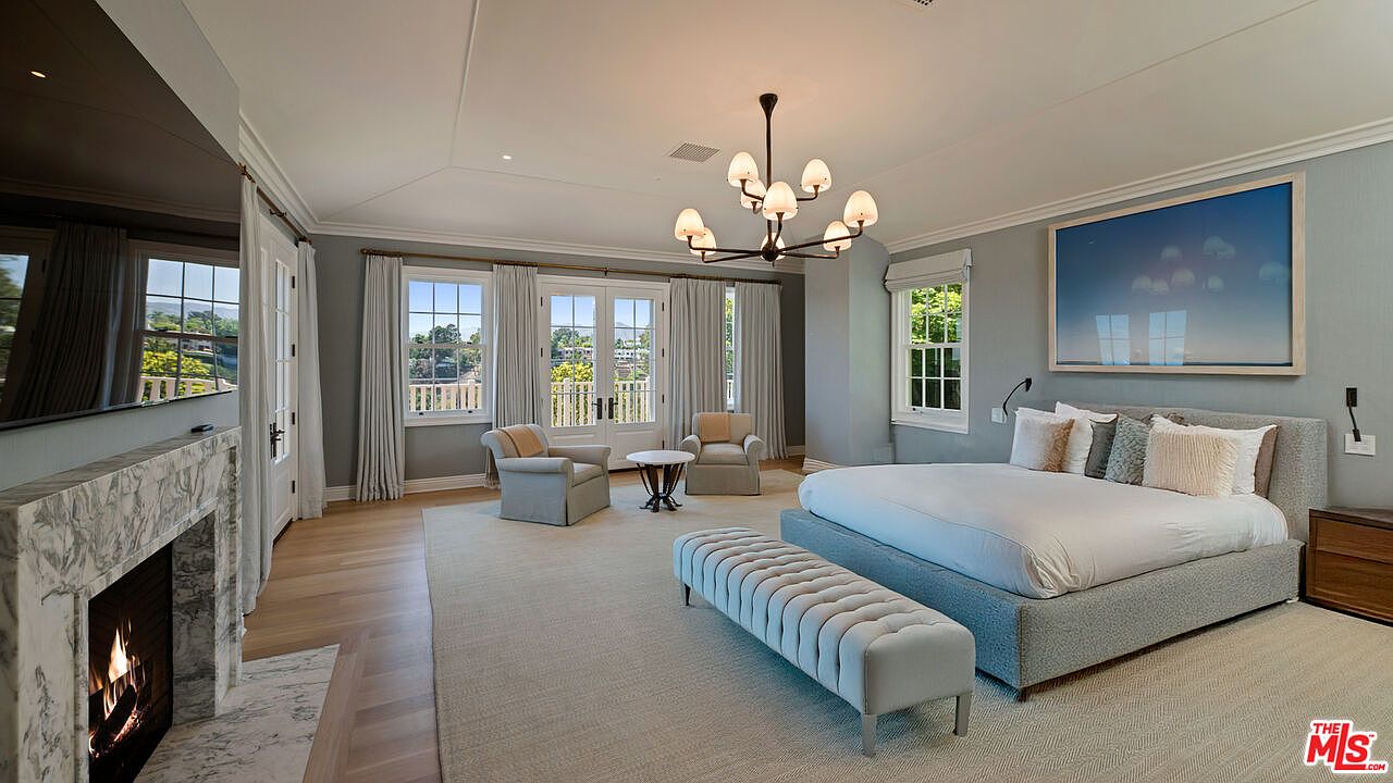 This is a spacious primary bedroom featuring a large bed with a tufted headboard, a seating area with two armchairs and a small table, and a fireplace with a marble surround. The room is decorated in neutral tones with gray walls and light-colored flooring, creating a serene and luxurious atmosphere. Natural light floods the room through multiple windows and French doors, enhancing the sense of openness and comfort.