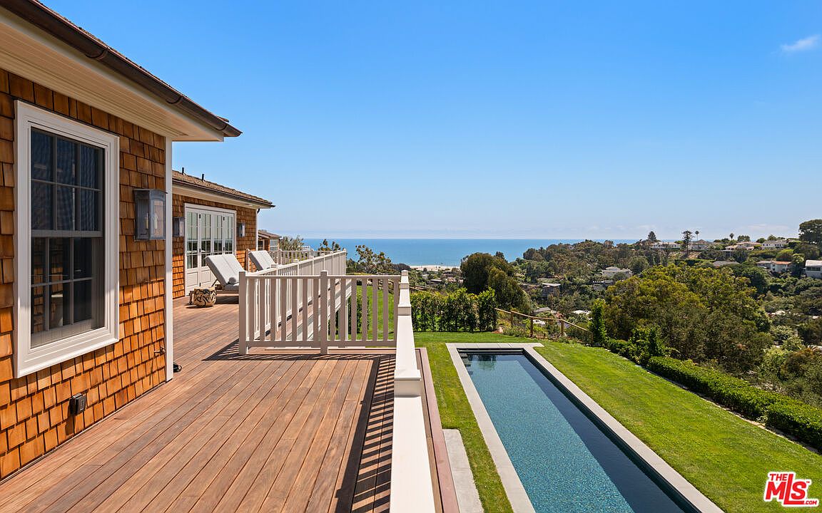 This image showcases a beautiful outdoor deck and patio area with a stunning ocean view. The deck is made of wood and features a white railing, leading to a modern, narrow pool surrounded by lush green grass. The house has a classic shingle siding, and the overall impression is one of luxury and tranquility.