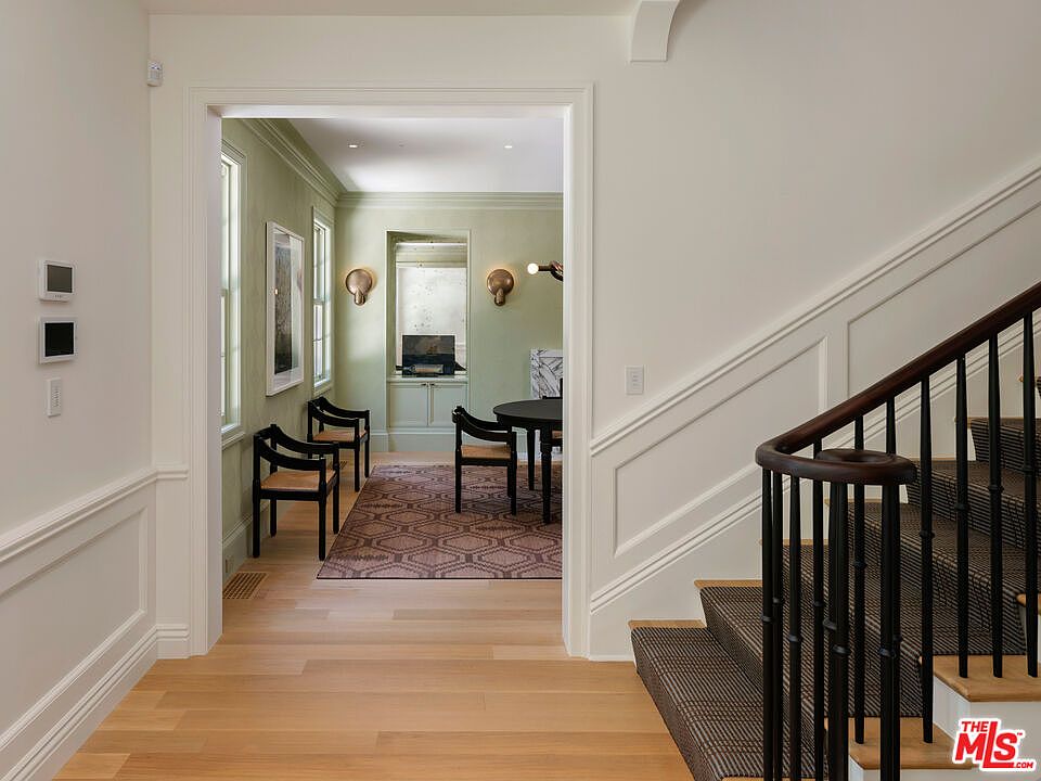 This interior shot showcases a hallway leading into a dining area, with a staircase on the right. The hallway features light hardwood flooring and white wainscoting, while the dining area has a patterned rug and green walls. The staircase has a dark wood railing and carpeted steps, creating a transition between spaces.