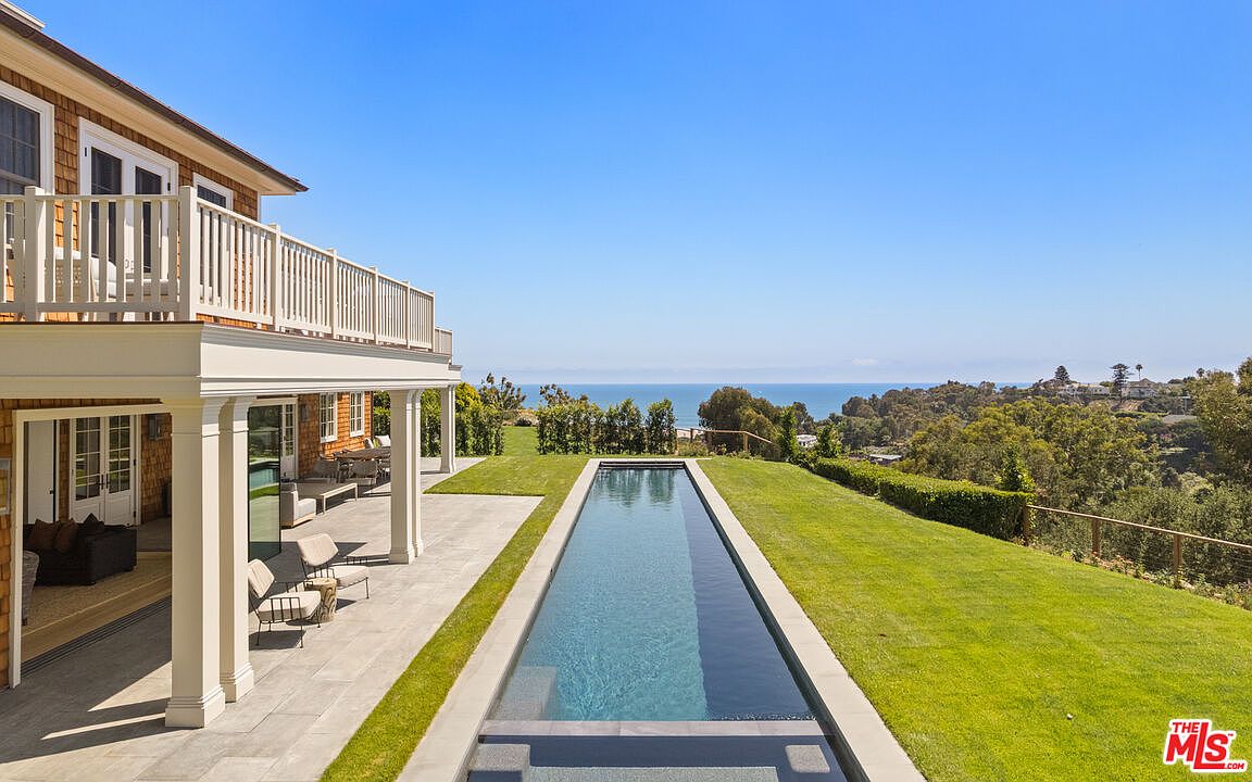 This image showcases a stunning backyard with a long, rectangular pool that appears to stretch towards the ocean view in the distance. The pool is surrounded by lush green grass and a stone patio area, with a portion of the house featuring a balcony visible on the left. The scene evokes a sense of luxury and relaxation, highlighting the property's outdoor living space and scenic location.