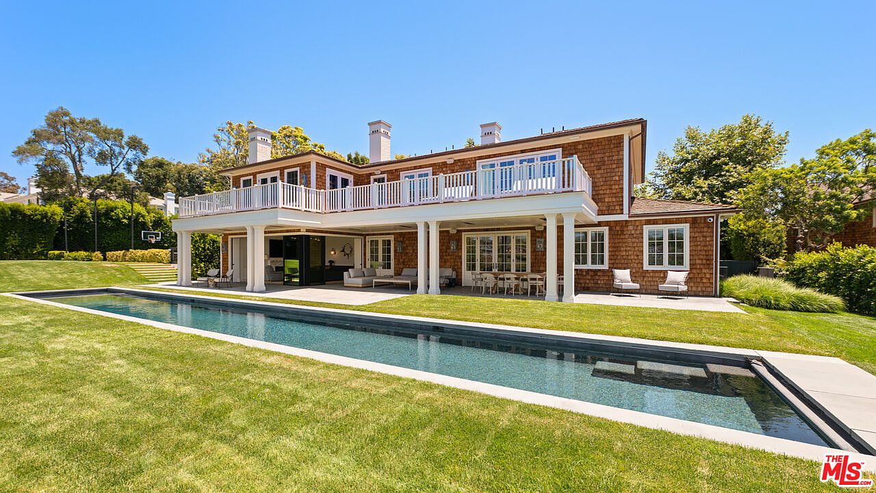 This image showcases the rear exterior of a luxurious home, highlighting a long, narrow swimming pool that stretches across the well-manicured lawn. The house features a two-story structure with a balcony, cedar shake siding, and white columns supporting the covered patio area. Outdoor seating and dining arrangements are visible, suggesting an ideal space for relaxation and entertainment.