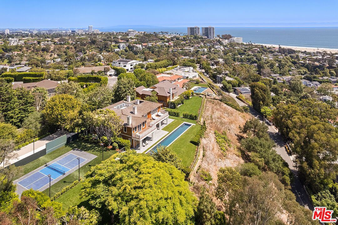 This aerial shot showcases a luxurious estate with a sprawling layout, featuring a large house with balconies, a long swimming pool, and a tennis court. The property is surrounded by lush greenery and mature trees, offering privacy and a sense of seclusion. In the background, the ocean and city skyline provide a stunning backdrop, emphasizing the prime location and upscale ambiance.