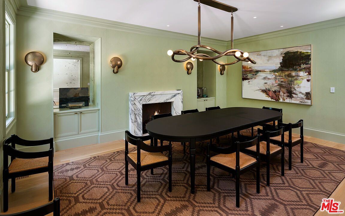 This dining room features a long, black oval table surrounded by black chairs with woven seats, set upon a patterned rug. A modern, sculptural chandelier hangs above the table, and a marble fireplace adds a touch of elegance. The walls are painted a soft green, and a large abstract painting hangs on the wall, creating a sophisticated and inviting atmosphere.