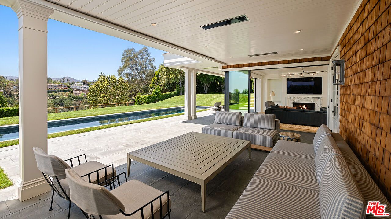 This image showcases a luxurious outdoor patio area, seamlessly blending indoor and outdoor living. The space features comfortable lounge seating, a large coffee table, and a view of a pristine pool and manicured lawn. The design incorporates natural wood elements and a neutral color palette, creating a sophisticated and relaxing atmosphere.
