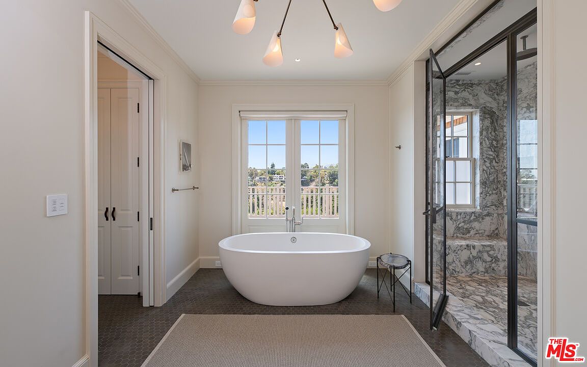 This is a bright and airy primary bathroom featuring a freestanding white bathtub positioned in front of a large window with a view. The bathroom has a modern aesthetic with a glass-enclosed shower, dark-toned hexagonal floor tiles, and a neutral-colored rug. A stylish light fixture hangs from the ceiling, adding to the room's elegant ambiance.