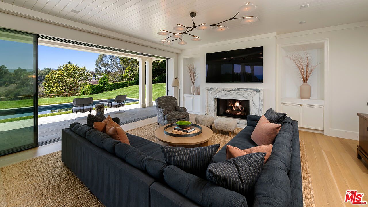 This is a bright and airy living room featuring a large sectional sofa, a round coffee table, and a fireplace with a television mounted above. The room opens to an outdoor patio and pool area through large sliding glass doors, creating a seamless indoor-outdoor living experience. The neutral color palette and natural materials contribute to a sophisticated and inviting atmosphere.