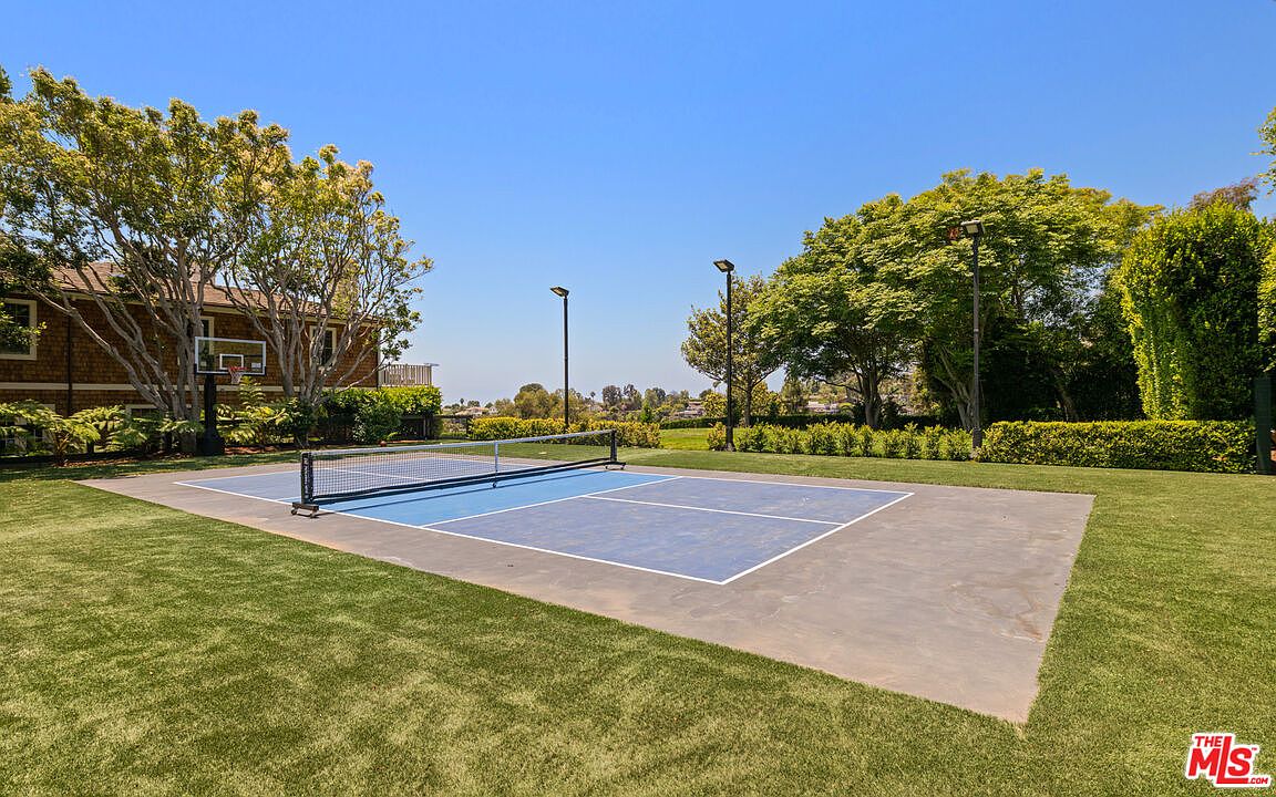 This image showcases a well-maintained yard featuring a blue and gray sports court, possibly for tennis or pickleball, surrounded by lush green grass. Mature trees and manicured hedges frame the area, providing privacy and a sense of tranquility. A basketball hoop is visible near a building in the background, suggesting recreational opportunities.