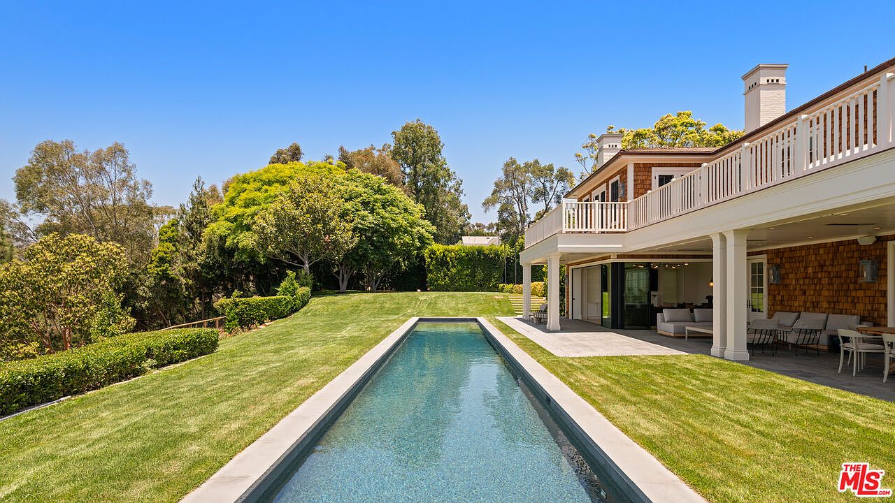 This image showcases a luxurious backyard featuring a long, rectangular swimming pool with a light-colored stone border. The pool is surrounded by a well-manicured lawn and lush greenery, leading up to a two-story house with a balcony and outdoor seating area. The scene exudes a sense of tranquility and upscale outdoor living.