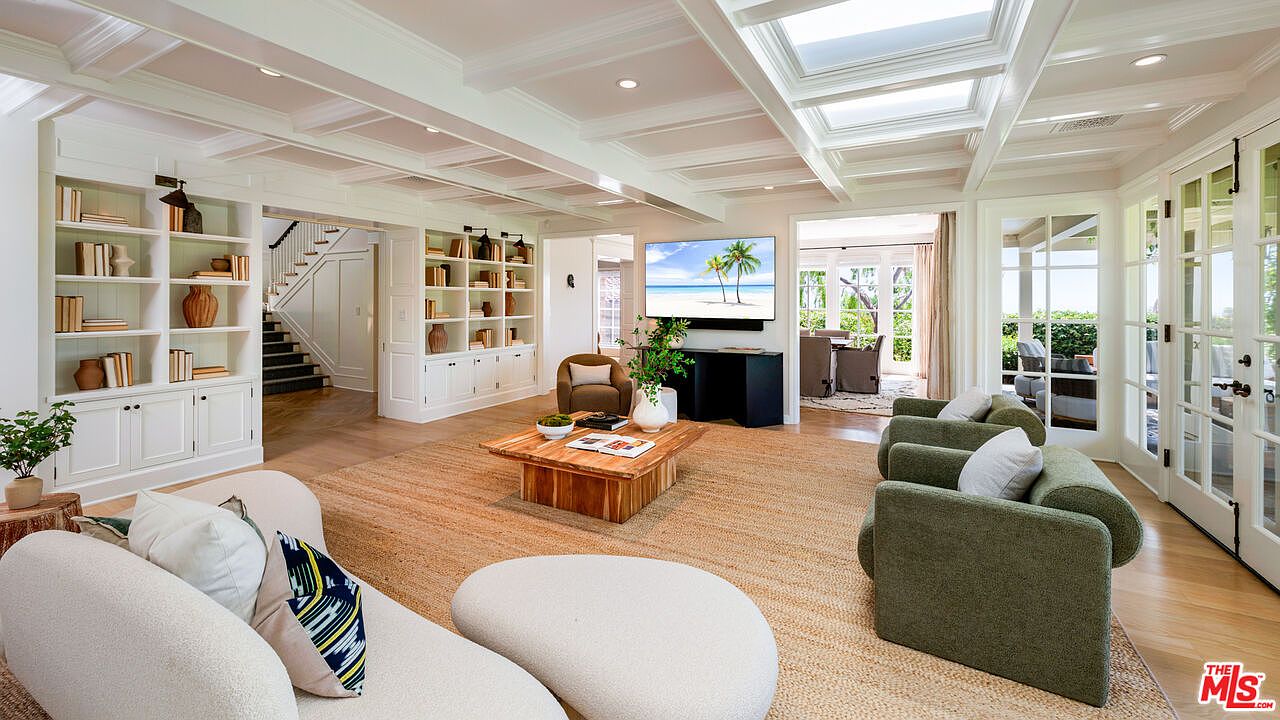 This is a bright and spacious living room featuring built-in bookshelves, a coffered ceiling with skylights, and hardwood floors covered by a large area rug. The room is furnished with modern sofas and chairs, and a large television is mounted above a console. French doors lead to an outdoor area, enhancing the indoor-outdoor flow.