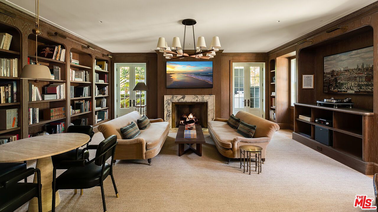 This is an interior shot of a living room featuring dark wood paneling and built-in bookshelves. Two sofas face each other in front of a fireplace with a TV mounted above it displaying a beach scene. A round table with chairs is visible on the left, and the room is illuminated by a modern chandelier, creating a warm and inviting atmosphere.
