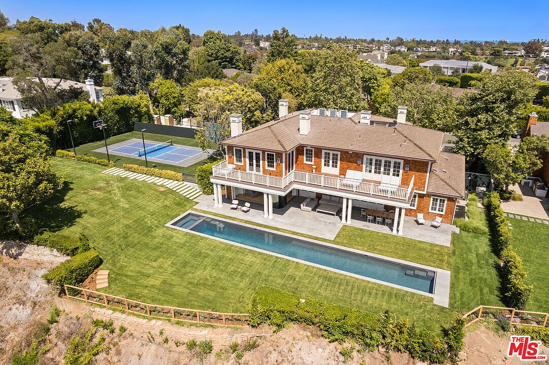 This aerial shot showcases a luxurious home with a well-manicured lawn, a long swimming pool, and a tennis court. The house features a classic design with a brown roof and white trim, complemented by lush greenery and mature trees. The overall impression is one of elegance and privacy.