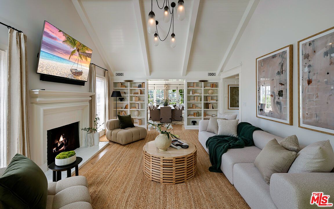 This is a bright and airy living room featuring a vaulted ceiling with exposed beams, built-in bookshelves, and a cozy fireplace. The room is furnished with a neutral-toned sofa, a round coffee table, and a comfortable armchair, creating an inviting and sophisticated atmosphere. A large television is mounted above the fireplace, and natural light floods the space through the windows.