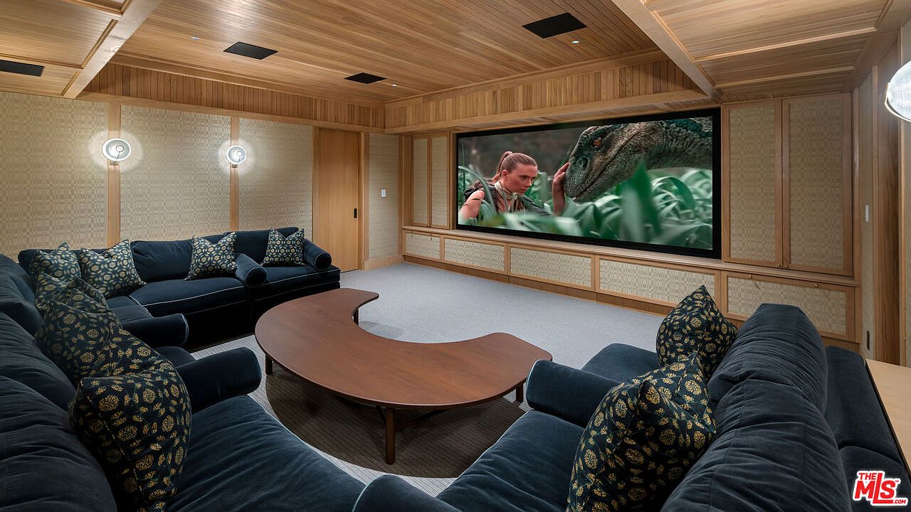 This is a home theater room featuring a large sectional sofa in a dark blue velvet fabric with patterned throw pillows. A curved wooden coffee table sits in the center of the room, facing a large screen displaying a movie. The walls and ceiling are clad in wood paneling, creating a warm and inviting atmosphere.
