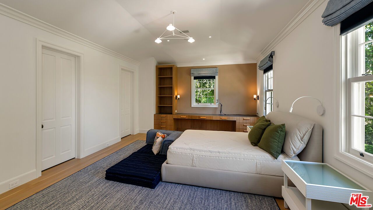 This is a well-lit bedroom featuring a bed with a light-colored mattress and headboard, complemented by olive green pillows. A built-in desk and shelving unit provide ample storage and workspace. The room is decorated with a blue rug and a modern light fixture, creating a comfortable and stylish atmosphere.