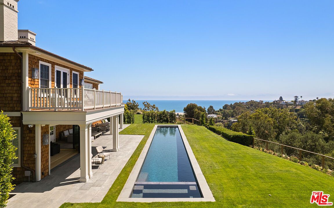 This image showcases a luxurious backyard featuring a long, rectangular swimming pool with a view of the ocean. The pool is surrounded by a well-manicured lawn, and the property includes a house with a balcony overlooking the pool area. The scene evokes a sense of tranquility and high-end living.