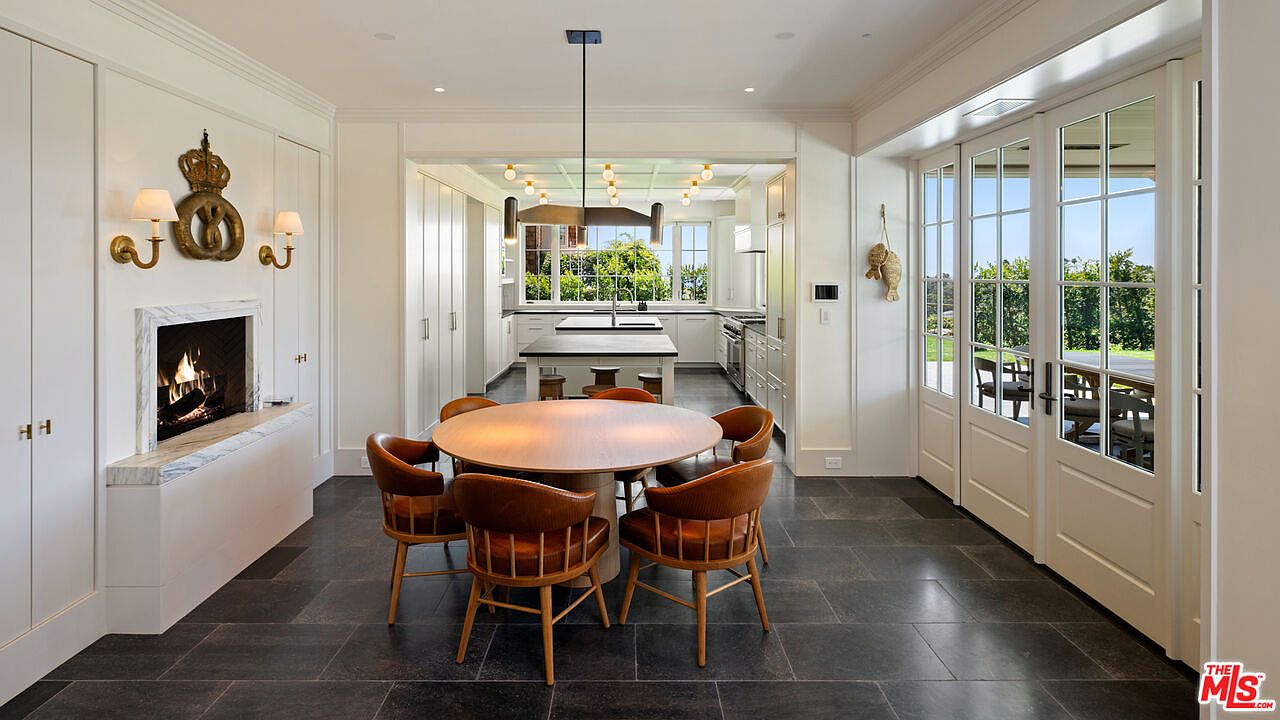 This is an interior shot of a dining room that seamlessly transitions into a kitchen area. The dining room features a round wooden table surrounded by six leather chairs, set upon dark stone flooring. A fireplace adds a cozy touch, while large glass doors offer views of the outdoor greenery, creating a bright and inviting space.