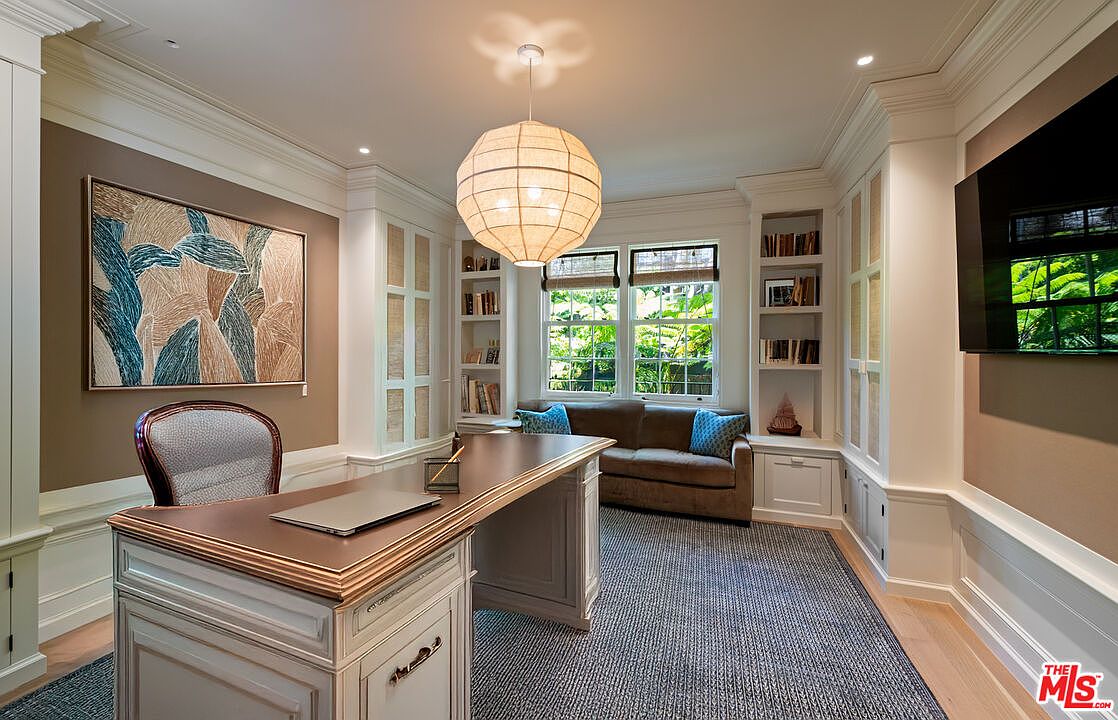 This is an interior shot of a well-appointed home office. The room features custom built-in shelving and cabinetry, a large desk with a laptop, and a comfortable sofa beneath a window. A modern light fixture hangs from the ceiling, and artwork adorns the walls, creating a sophisticated and functional workspace.