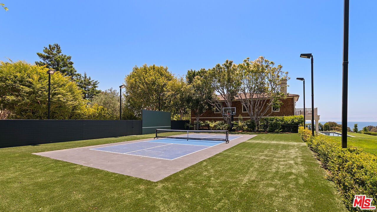 This image showcases a well-maintained yard featuring a pickleball court with a blue and gray surface. The court is surrounded by lush green grass and manicured hedges, with trees and a building visible in the background. The scene is bathed in sunlight, creating an inviting and recreational outdoor space.
