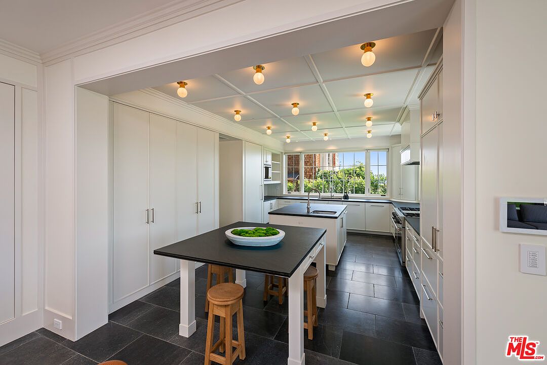 This is a bright and spacious kitchen featuring white cabinetry, dark countertops, and a dark tile floor. A central island with a black countertop and wooden stools provides a gathering space, while recessed lighting illuminates the coffered ceiling. The kitchen is well-lit with natural light from a large window overlooking the backyard.