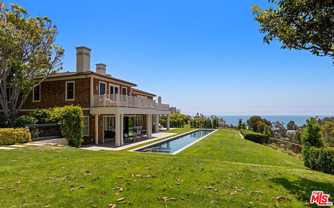 This image showcases the rear exterior of a luxurious home, featuring a shingle-style facade, a spacious balcony, and a long, rectangular pool seamlessly integrated into the manicured lawn. The property overlooks a stunning ocean view, enhancing its appeal as a high-end real estate offering. The perspective is from a low angle, emphasizing the size of the house and the depth of the yard.