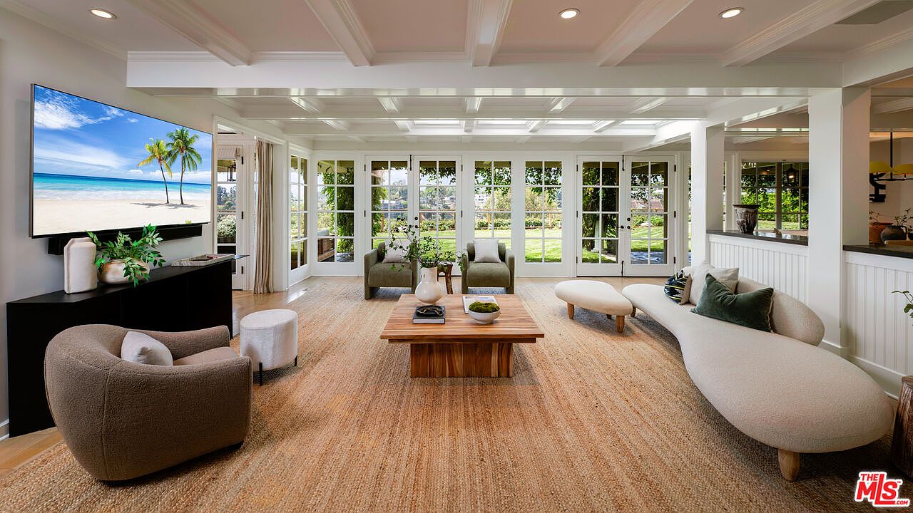 This is a bright and airy living room featuring a large flat-screen TV displaying a beach scene, a modern curved sofa, and two armchairs. The room has multiple French doors leading to the outside, allowing for ample natural light and views of the garden. The coffered ceiling and neutral color palette create a sophisticated and inviting atmosphere.