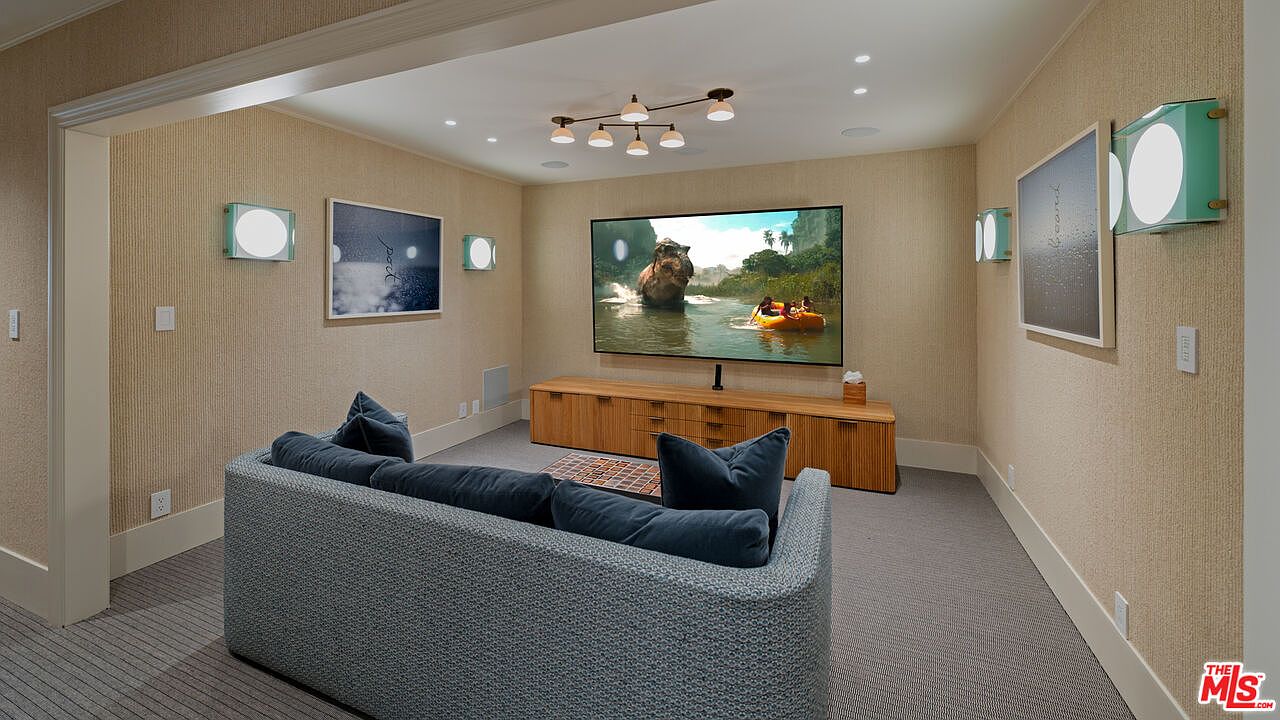This is an interior shot of a living room or media room, featuring a large television displaying a scene with people in a raft, a modern sofa with blue cushions, and a wooden media console. The room is decorated with framed artwork and unique wall sconces, creating a cozy and stylish entertainment space. The walls are covered in a textured wallpaper, and the lighting includes recessed lights and a modern chandelier.