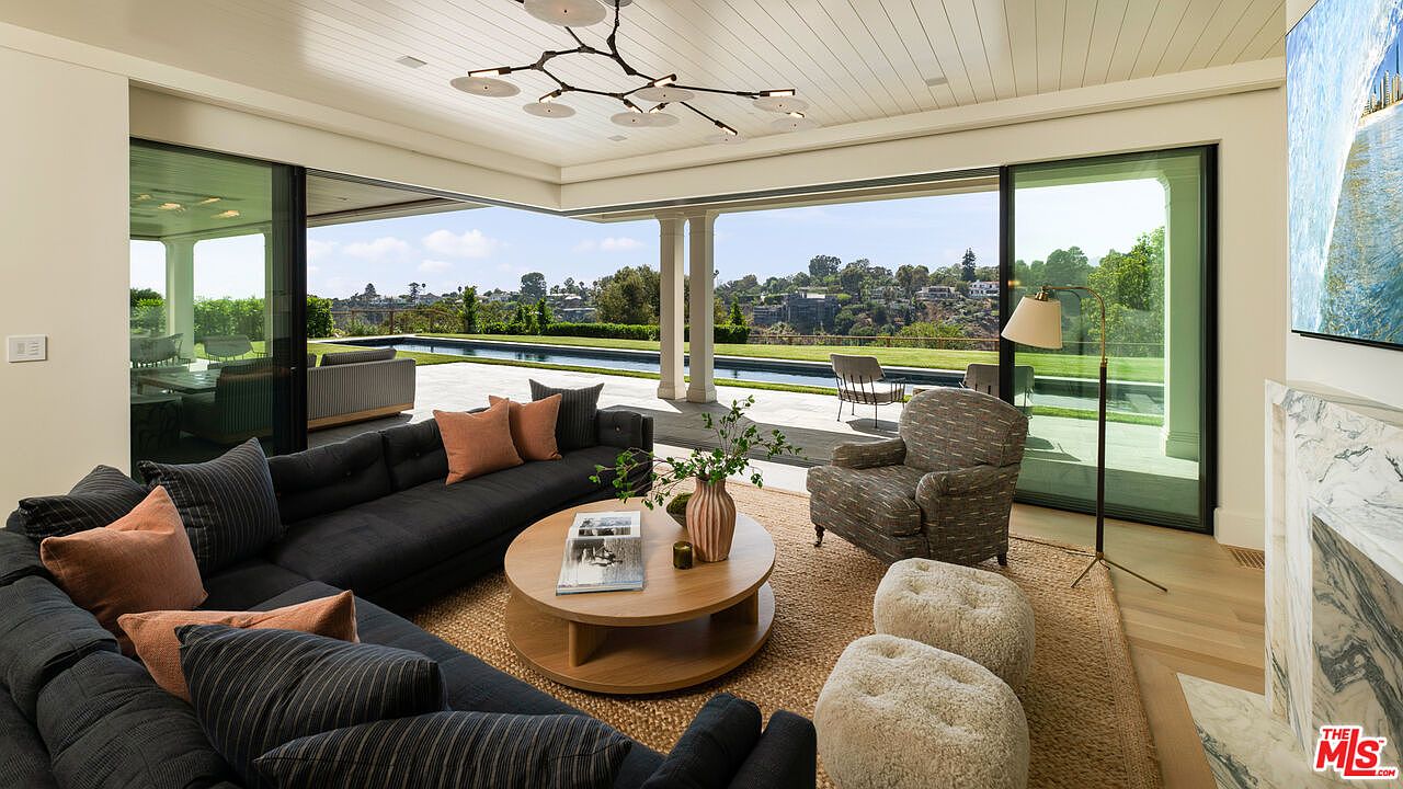 This is a beautifully staged living room with an open floor plan that seamlessly blends indoor and outdoor living. The room features a large sectional sofa, a round coffee table, and a stylish armchair, all arranged on a jute rug. Sliding glass doors open to a patio with a pool and landscaped grounds, offering stunning views and creating a bright and airy atmosphere.