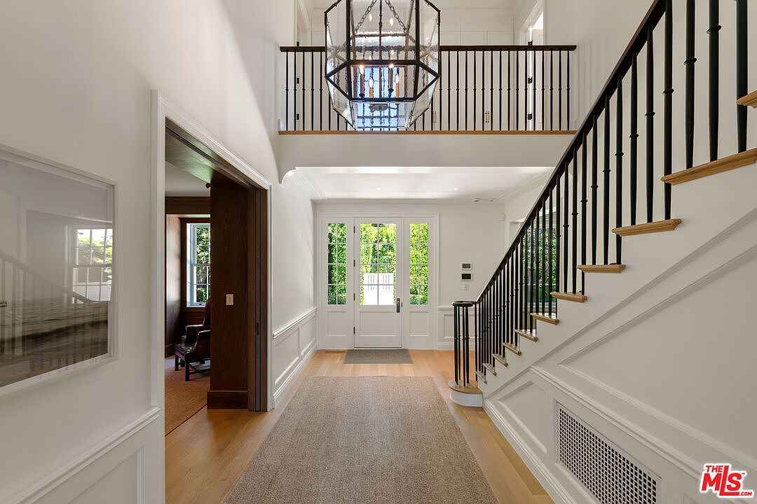 This interior shot showcases a grand hallway with a staircase. The space features white walls with wainscoting, hardwood floors, and a large area rug. A striking black railing contrasts with the light-colored stairs, and a modern chandelier hangs above the entryway, creating an elegant and inviting atmosphere.