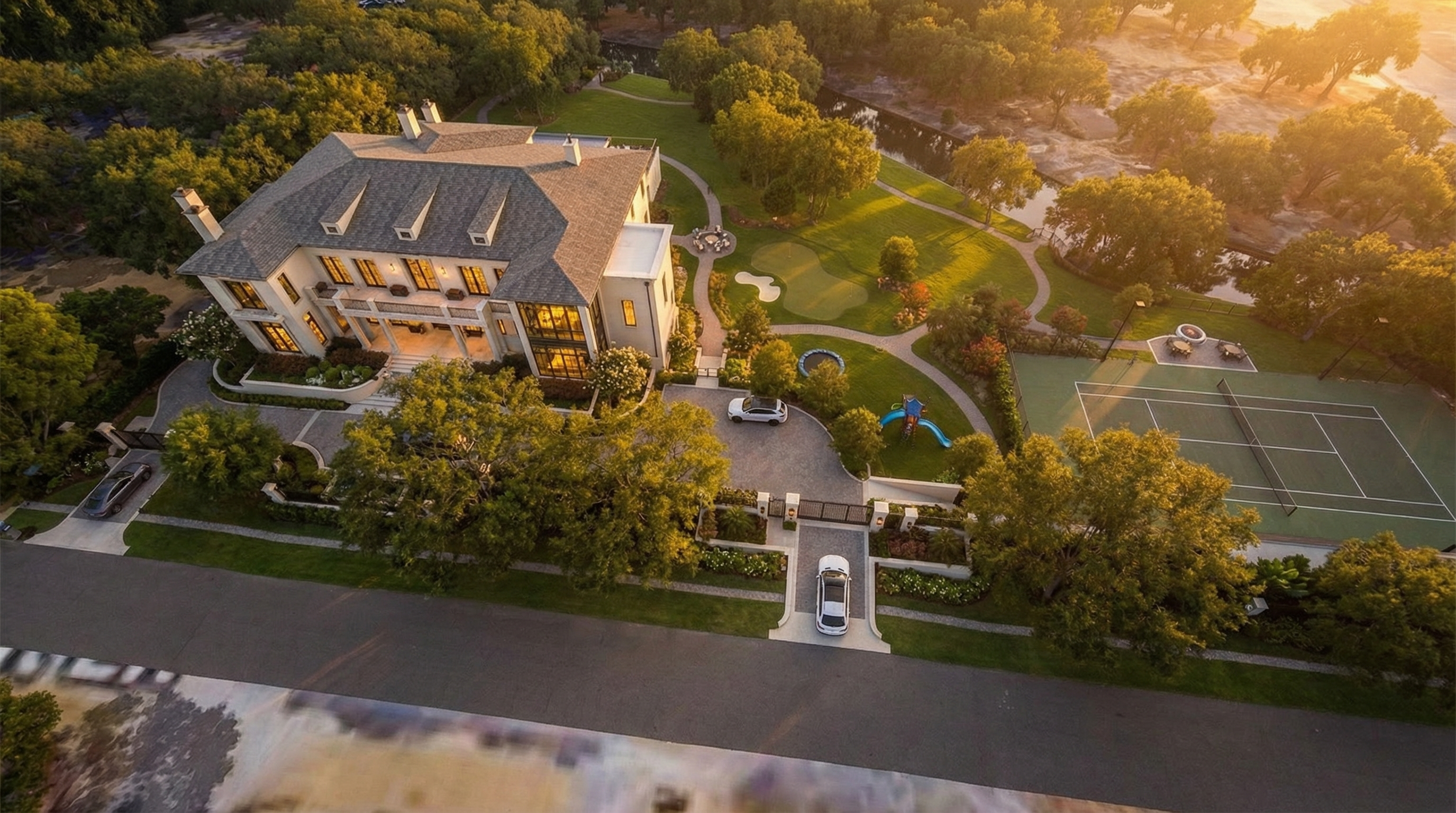 This aerial view showcases a grand estate with a meticulously landscaped yard, including a tennis court, putting green, and children's play area. The stately home features a symmetrical facade, multiple dormers, and a rooftop terrace, complemented by a circular driveway and mature trees. The property exudes luxury and offers a variety of outdoor amenities for recreation and relaxation.