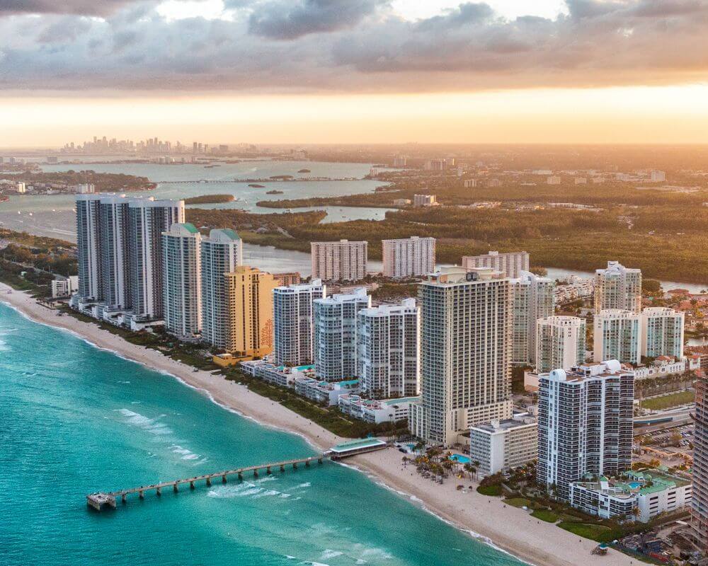 This aerial shot showcases a stunning coastal real estate development, demonstrating prime oceanfront property. Numerous high-rise condos line the beach, offering breathtaking views and direct access to the turquoise waters. Amenities like a pier and manicured lawns suggest a luxury lifestyle.