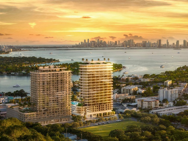 This aerial view showcases two modern high-rise residential buildings against a backdrop of a city skyline and ocean. The buildings feature sleek, contemporary architecture with multiple balconies. Below, lush greenery surrounds the property, suggesting a well-landscaped environment. The overall impression is one of luxury and urban living with scenic views.