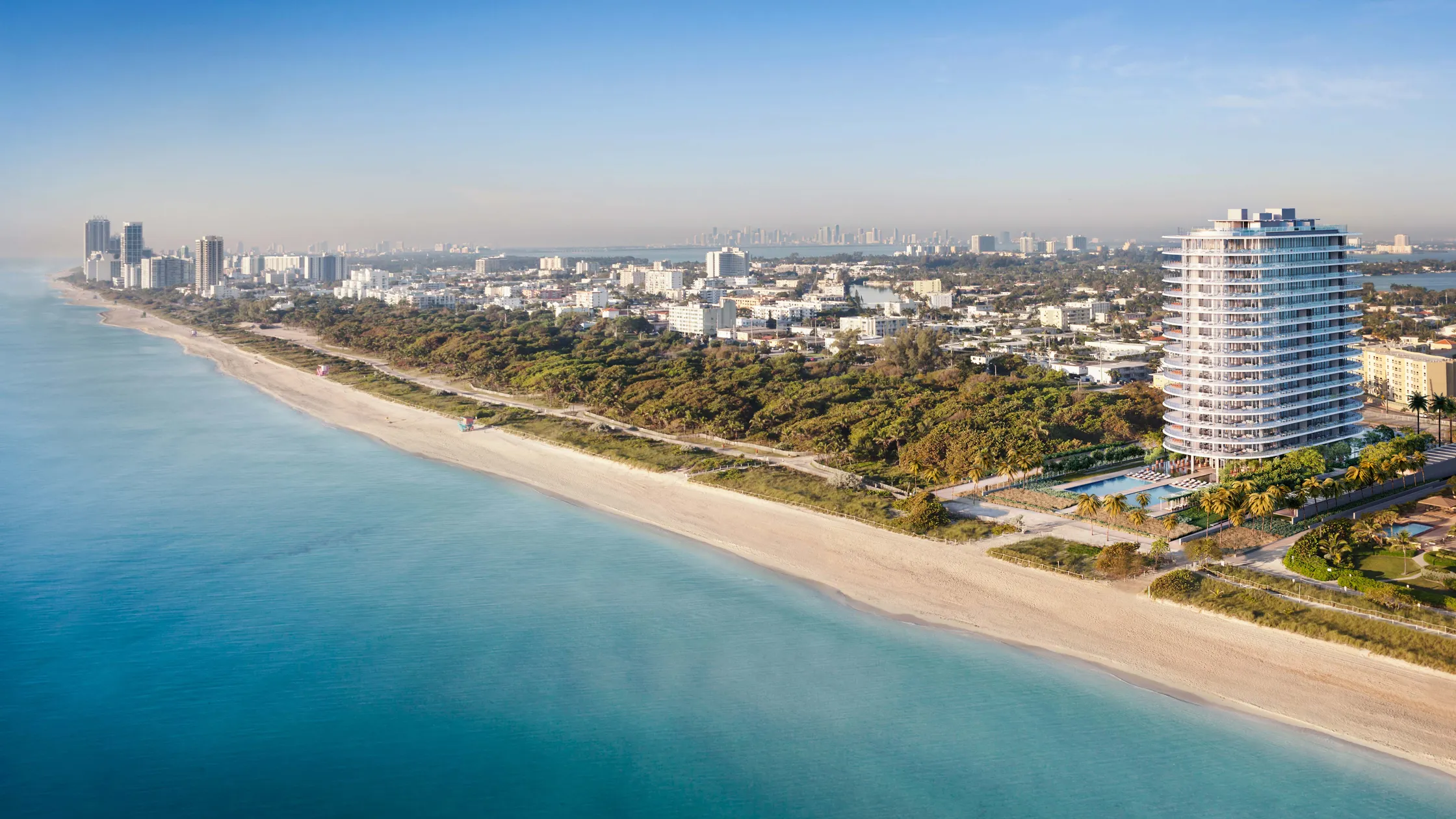 This aerial shot showcases an upscale beachfront property, highlighting a modern high-rise building with a distinctive rounded design. The building is situated near a pristine beach with turquoise waters, and features amenities such as a pool and landscaped grounds. The image emphasizes the property's desirable coastal location and luxurious lifestyle.