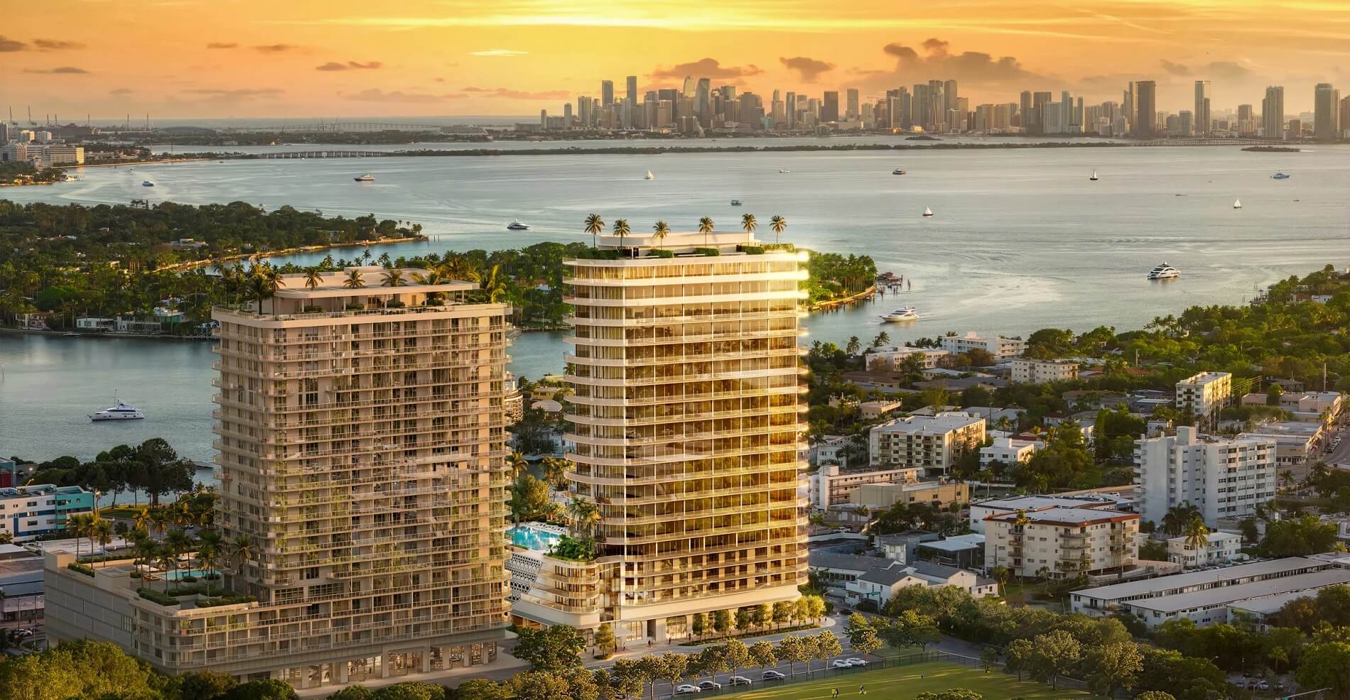 This aerial view showcases two modern high-rise residential buildings with sleek architectural designs, set against a backdrop of a bay and a distant city skyline at sunset. The buildings feature multiple balconies and rooftop amenities, including palm trees, suggesting luxury living. The surrounding area includes lush greenery and glimpses of residential neighborhoods, creating an impression of an upscale urban oasis.