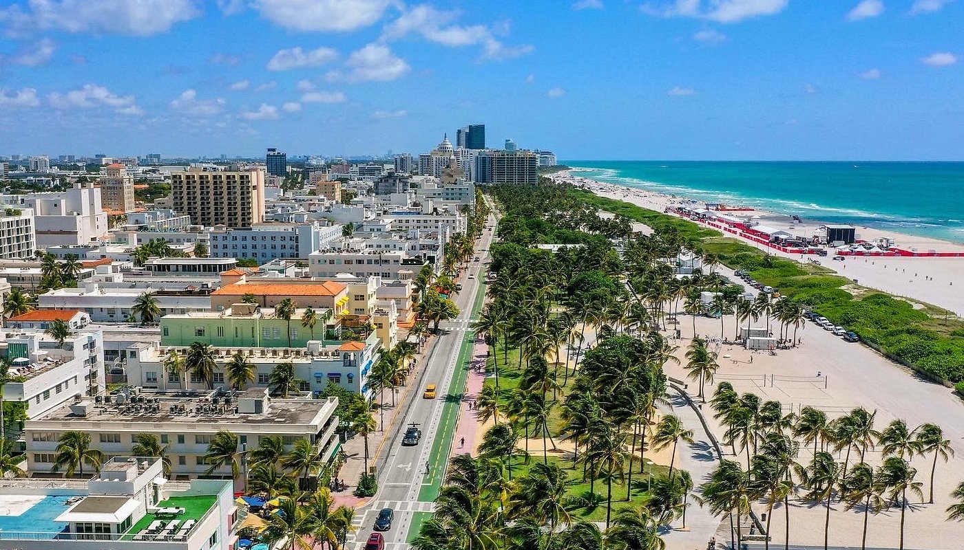 This is an aerial view of a beachfront cityscape. The image shows a mix of residential and commercial buildings, along with well-maintained streets lined with palm trees. The pristine beach and turquoise ocean create a sense of luxury and relaxation, ideal for highlighting the property's location and proximity to amenities.