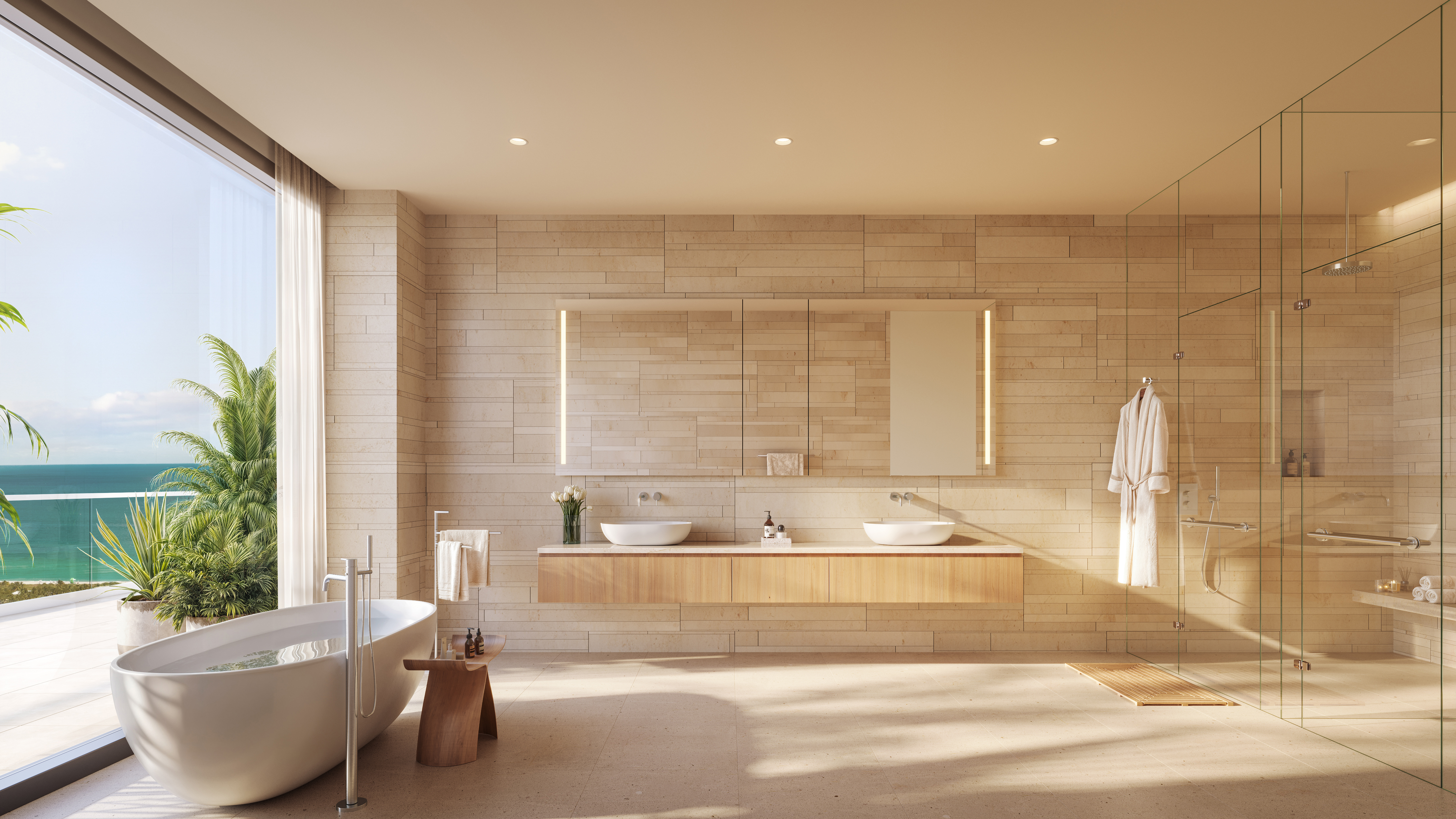 This is a luxurious primary bathroom featuring a double vanity with vessel sinks, a freestanding soaking tub next to a large window with ocean views, and a glass-enclosed shower. The walls are clad in light-colored stone tiles, and the overall design is modern and spa-like. The perspective is a wide shot, showcasing the entire space and its high-end finishes.