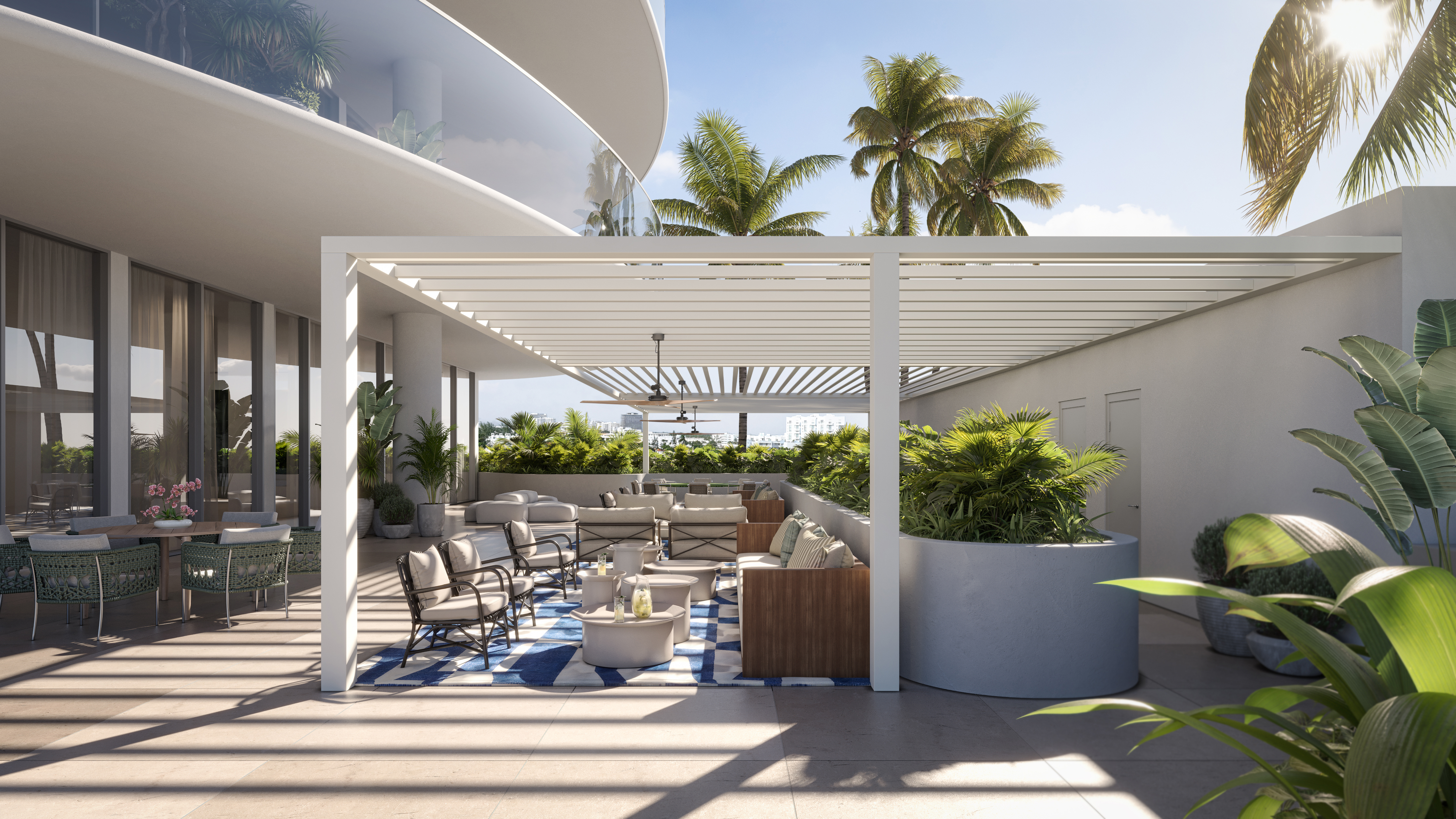 This image showcases a luxurious outdoor patio or balcony area, featuring stylish lounge furniture arranged on a blue and white patterned rug. The space is covered by a modern pergola, providing shade and architectural interest, while lush greenery in large planters adds a touch of nature. The overall impression is one of sophisticated outdoor living and relaxation.