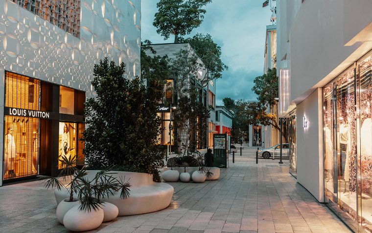This image showcases a high-end retail street with designer stores like Louis Vuitton and Cartier. The architecture is modern and luxurious, with sleek white buildings and thoughtfully designed outdoor spaces including unique seating areas and landscaping. The image projects an upscale, sophisticated ambiance suitable for highlighting the prime location of a real estate property.