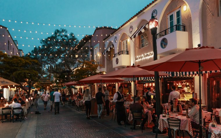 This image captures a vibrant outdoor dining area in a commercial district. The scene features tables set with checkered tablecloths under red umbrellas, suggesting a relaxed and inviting atmosphere. String lights overhead enhance the ambient lighting, making it an appealing space for evening dining and socializing.