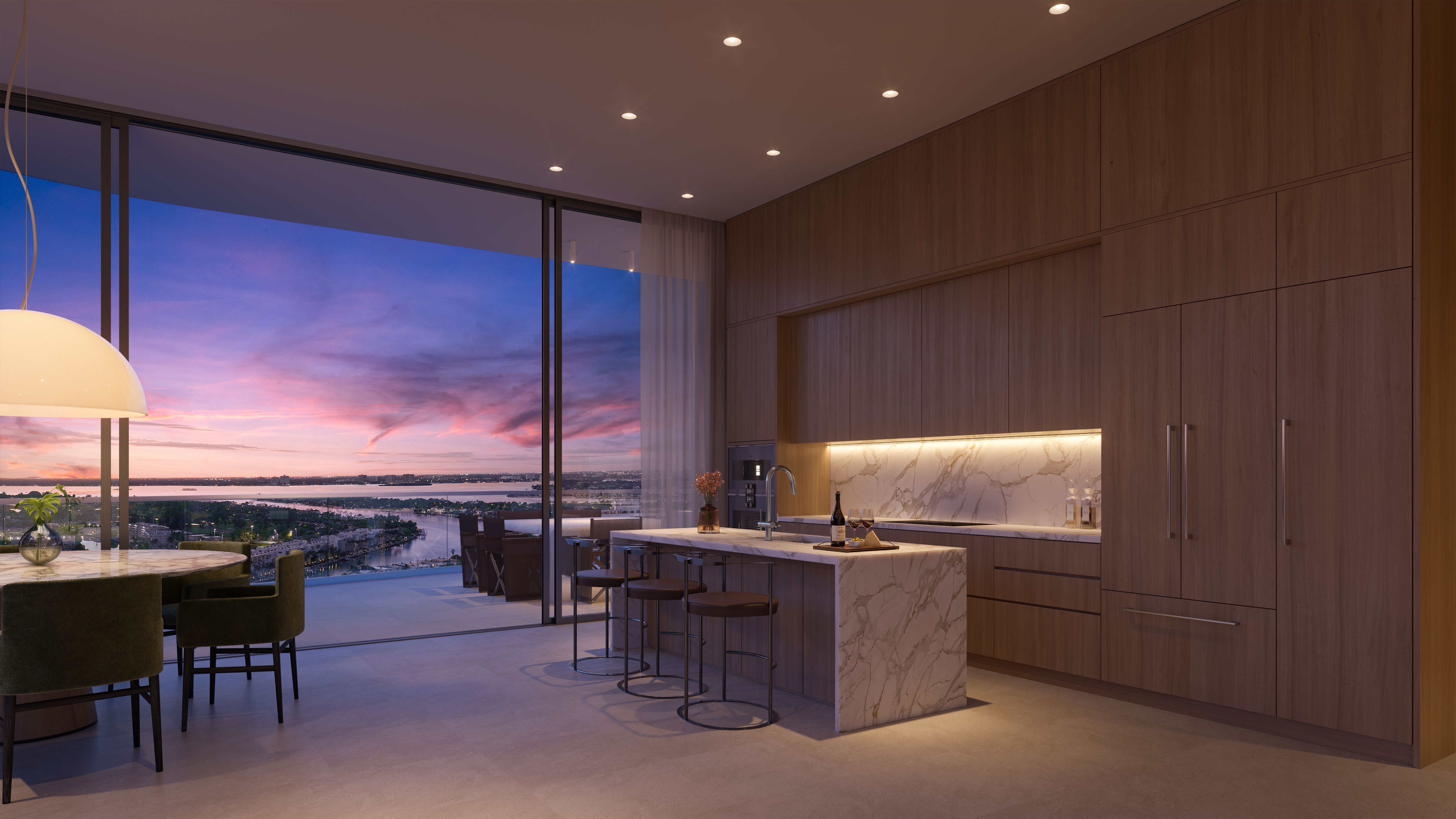 This is a modern kitchen with wood cabinetry, a marble island, and countertops. The kitchen features a large window with a view of the city and water at sunset. The open floor plan includes a dining area with a round table and green chairs.