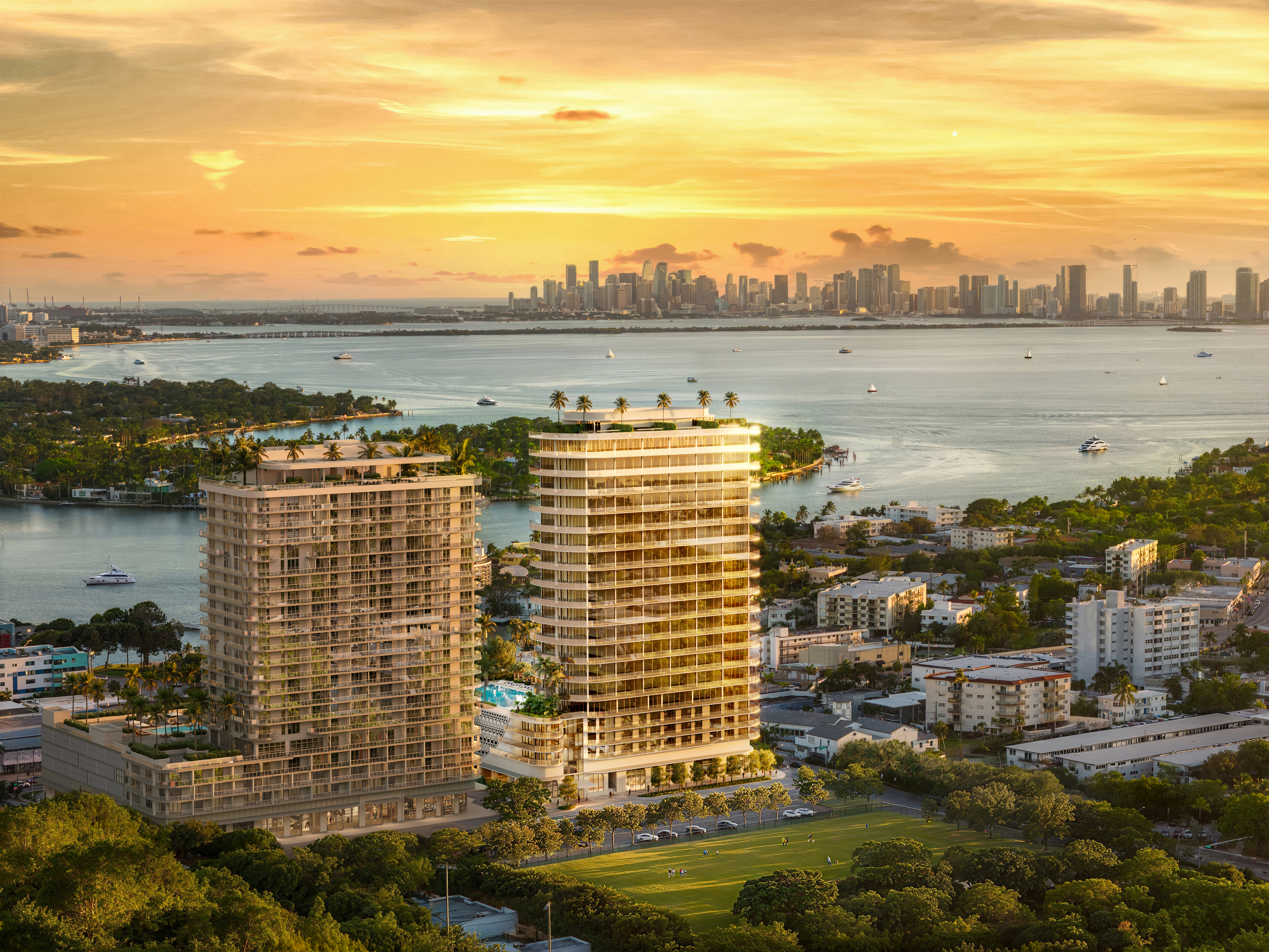 This aerial view showcases two modern residential towers with sleek architectural designs, complemented by lush greenery and a well-maintained green field in the foreground. The towers feature multiple balconies and rooftop amenities, while the cityscape and waterfront provide a stunning backdrop, creating an impression of luxury and urban living.