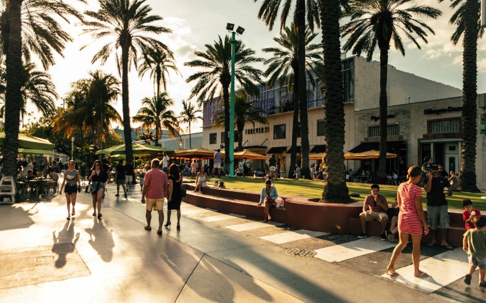 This image depicts an outdoor shopping area with palm trees, pedestrian walkways, and seating areas. Retail stores such as Forever 21 and Häagen-Dazs are visible in the background. The space creates a vibrant, walkable community atmosphere, showcasing amenities that add value to the surrounding real estate.